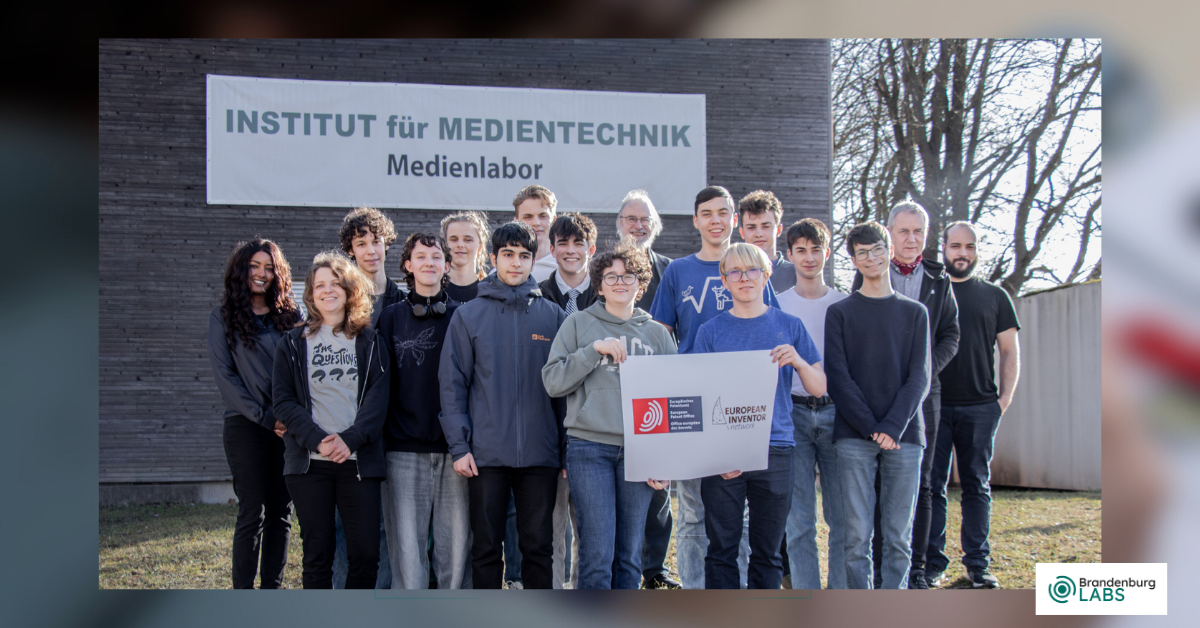 A group of students is standing with Prof. Karlheinz Brandenburg in front of the Institute of Media Technology at the Technical University of Ilmenau. Some of the students are holding a banner from the European Inventor Network.