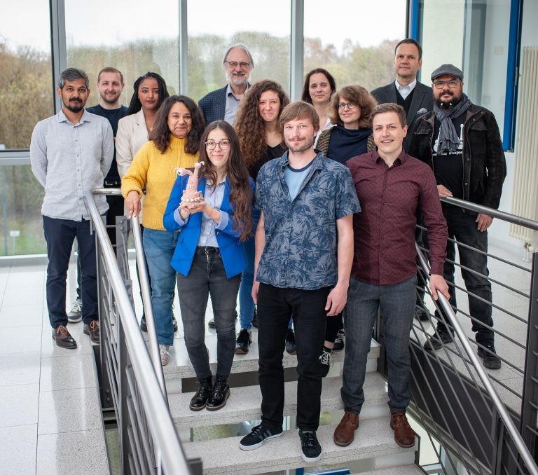 The Brandenburg Labs team including 13 team members is standing in the Technologie und Gründerzentrum Ilmenau. One woman holds the mascott , called Fribble in her hands.