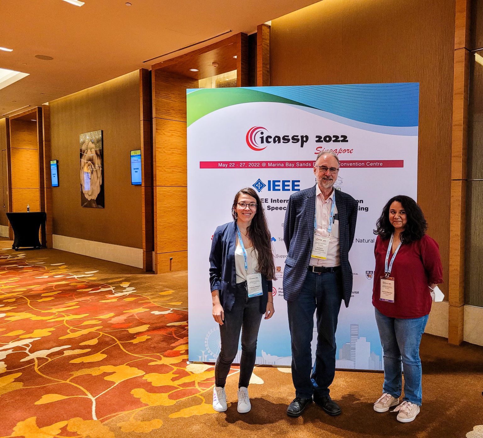Three people from Brandenburg Labs in front of a roll-up of the ICASSP 2022-